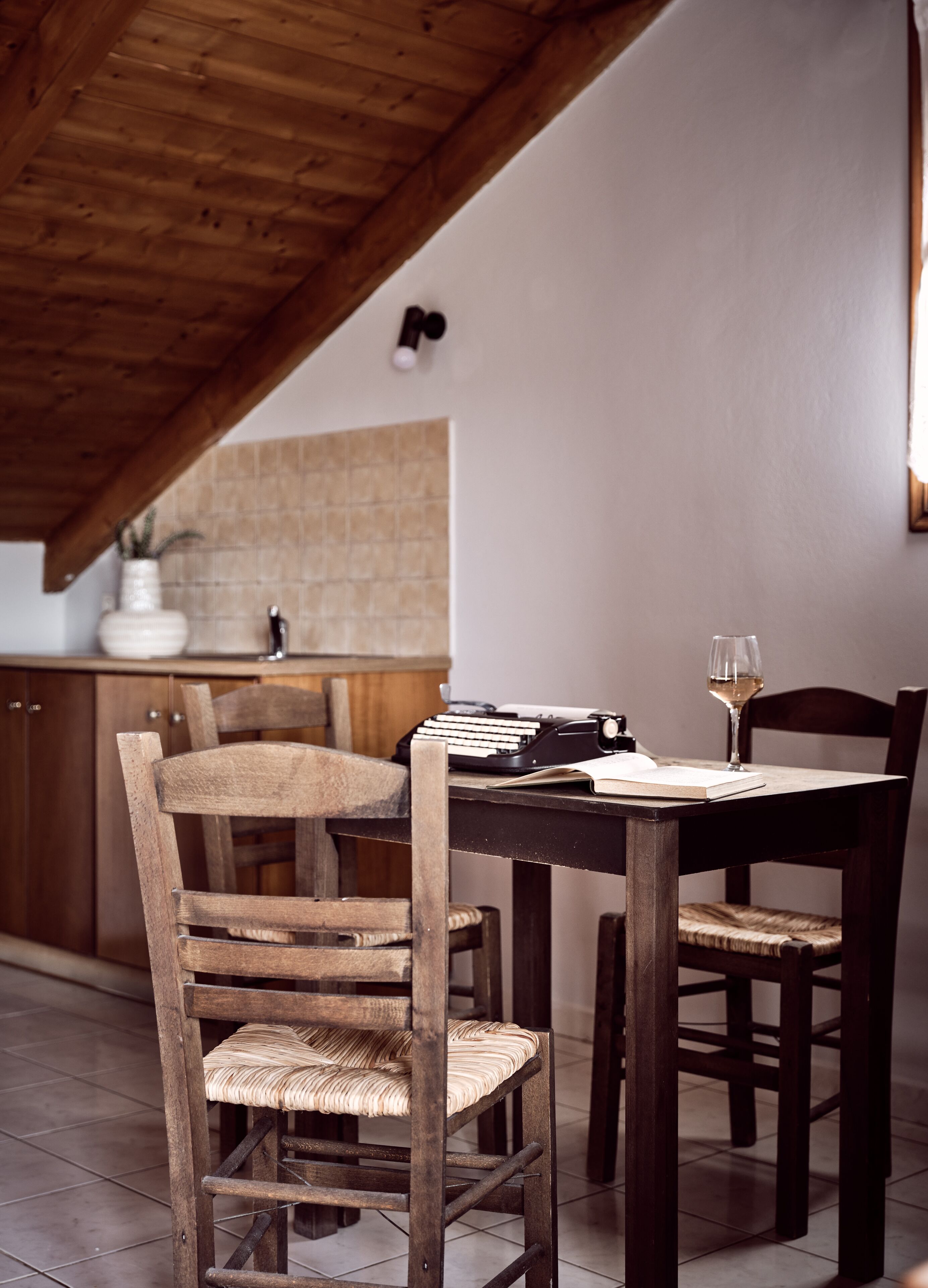 A dining area with wooden chairs and a table, a typewriter, and a wine glass.