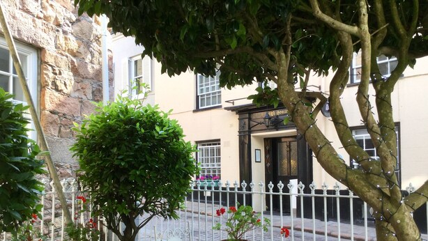 A courtyard with a stone wall, a white metal fence, and a table with two chairs. There are potted plants and a tree in the foreground. - Fabulous Apt Historic House St Aubin (St. Brelade)