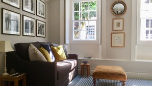 A living room with a dark brown sofa, multiple cushions, a small wooden side table, a blue and white striped rug, a round wooden ottoman, and framed artwork on the walls. - Fabulous Apt Historic House St Aubin (St. Brelade)