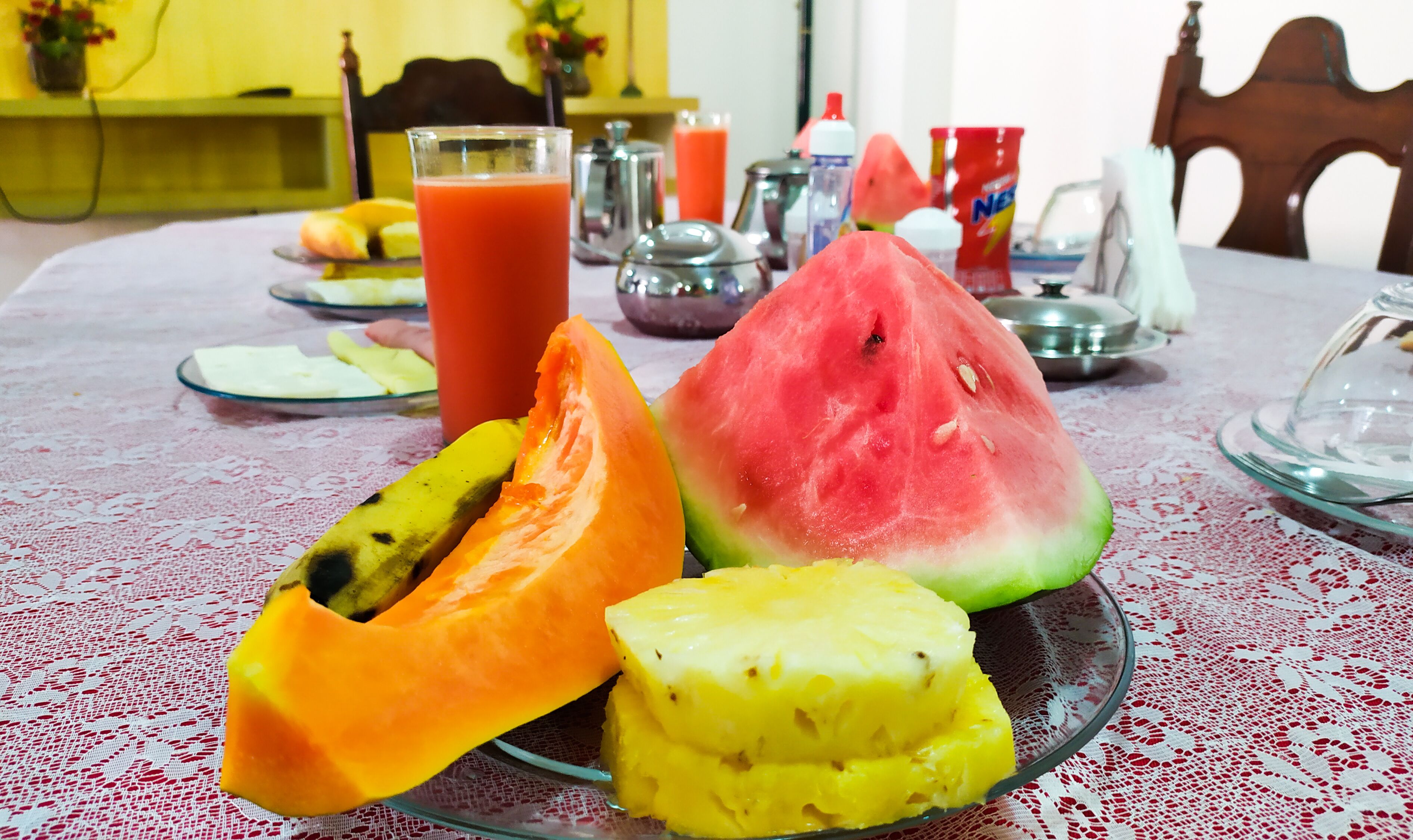 Una mesa con mantel rojo y blanco, un bol de vidrio con una tajada de sandía, un vaso de jugo de naranja y un vaso de jugo rojo.