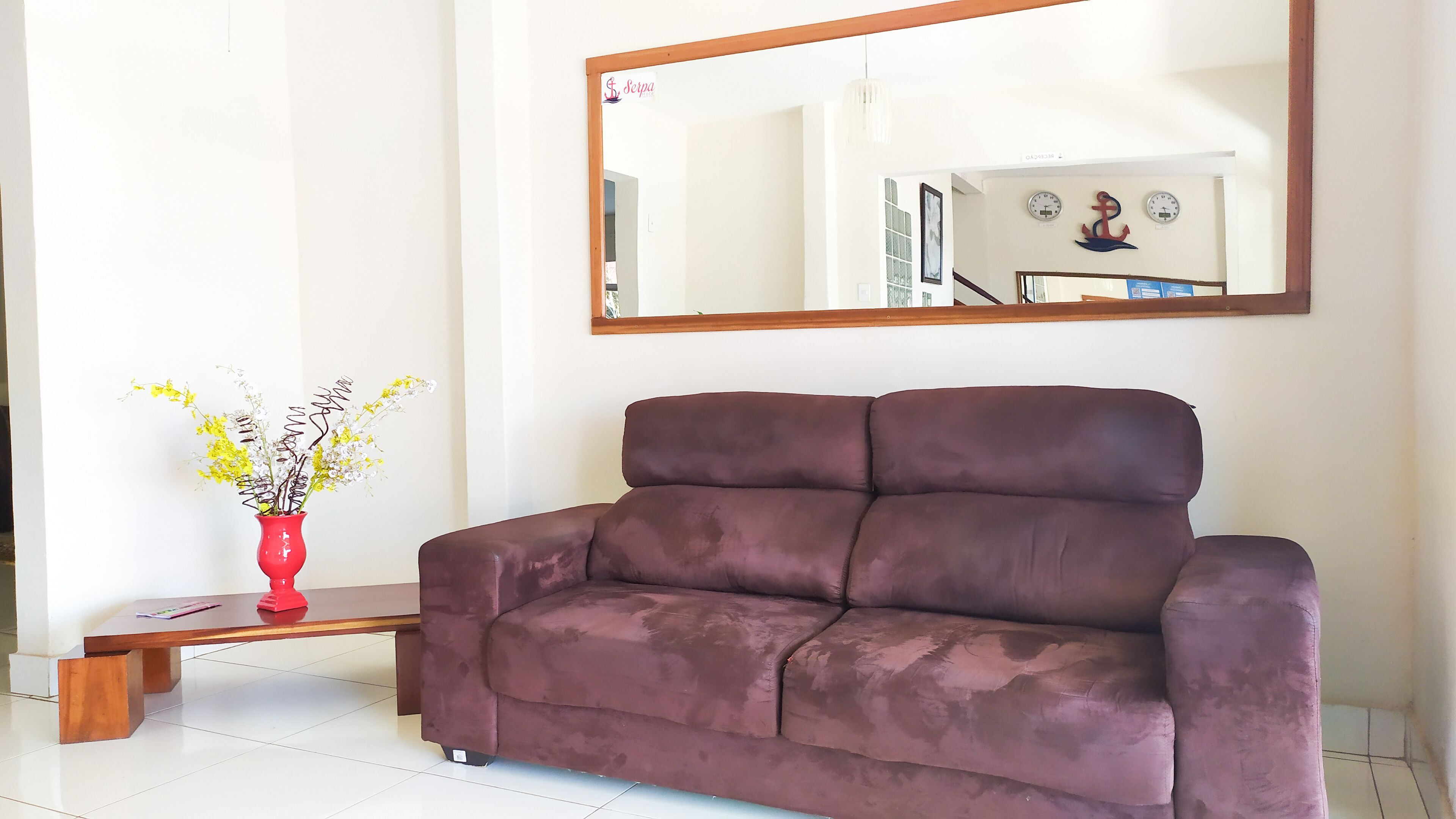 A brown sofa in a room with a large mirror on the wall, a red vase with flowers, and a wooden table.