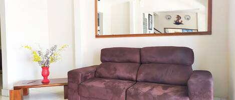 A brown sofa in a room with a large mirror on the wall, a red vase with flowers, and a wooden table.