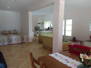 A room with a bed, a sofa, a dining table with a vase of flowers, and a kitchen area in the background.