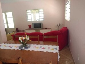 A small, cozy room with a wooden dining table, red upholstered chairs, a television on a white cabinet, and a window with white frames.