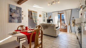 Un coin repas avec une table dressée pour deux personnes, un canapé et un espace cuisine équipé d’un réfrigérateur et d’une télévision.