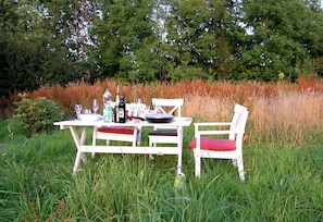 Une table extérieure blanche, accompagnée de chaises, entourée d’herbes hautes et d’arbres.