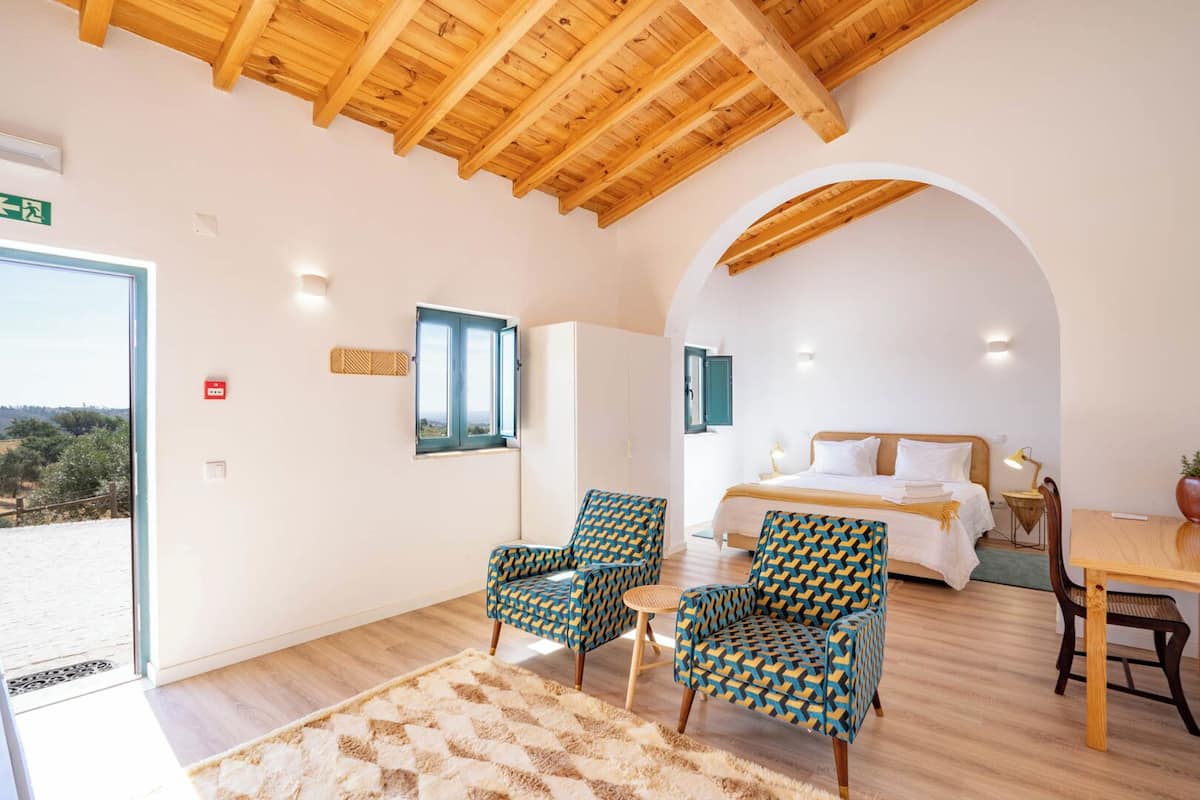 A room with a bed, two patterned armchairs, a small table, and a view of the outdoors.