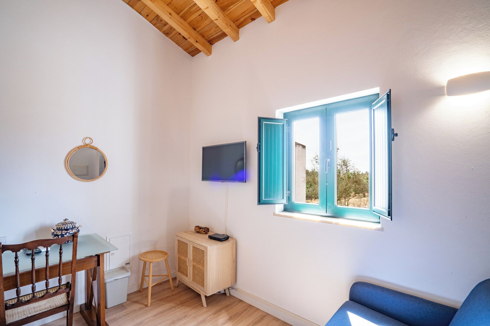 A room with a wooden ceiling, a TV mounted on the wall, a window with blue shutters, a wooden cabinet, and a dining table with chairs.