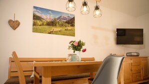 Ein Essbereich mit Holzzmöbeln, einer Vase mit Blumen, einem Gemälde von einer Berglandschaft und einem Fernsehgerät an der Wand.
