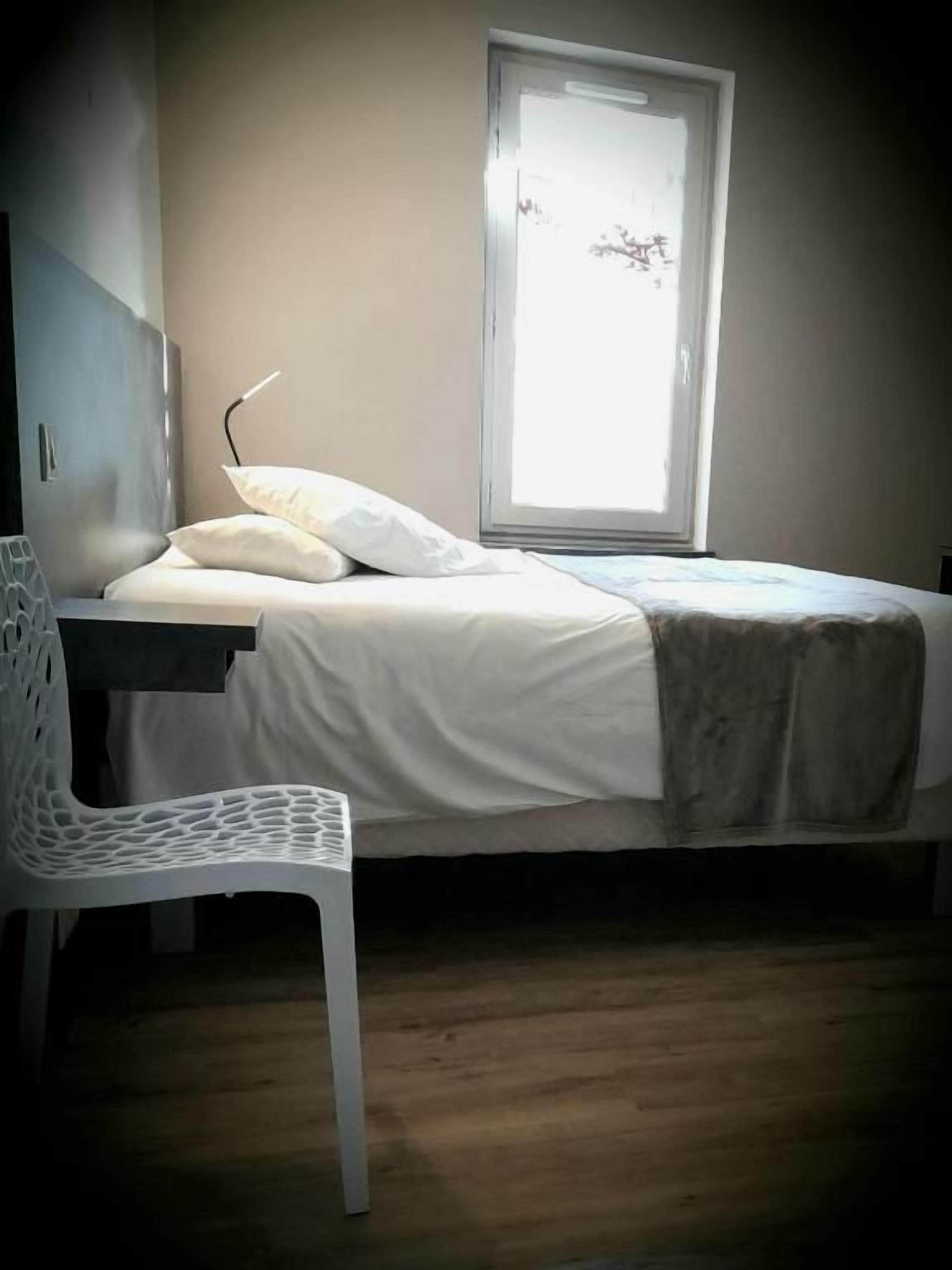 Una habitación pequeña con cama individual, ventana, una silla blanca y una mesita de noche.