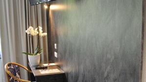 A modern room with a flat-screen TV mounted on the wall, a small wooden desk with a vase of flowers, a chair with a blue and white pattern, and a dark wood floor. - Hotel Restaurant Le Tout Va Bien (Valence)