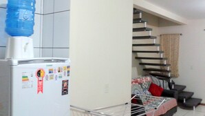 A compact living space with a white refrigerator, a water dispenser, a metal drying rack, a sofa with a floral cover, and a staircase. - Casa de Temporada Praia Santinho Inglese (Florianópolis)
