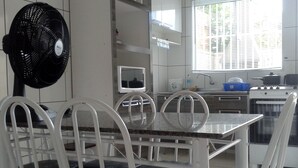 A dining area with a table and chairs, a black fan, a television, and a window with blinds. - Casa de Temporada Praia Santinho Inglese (Florianópolis)