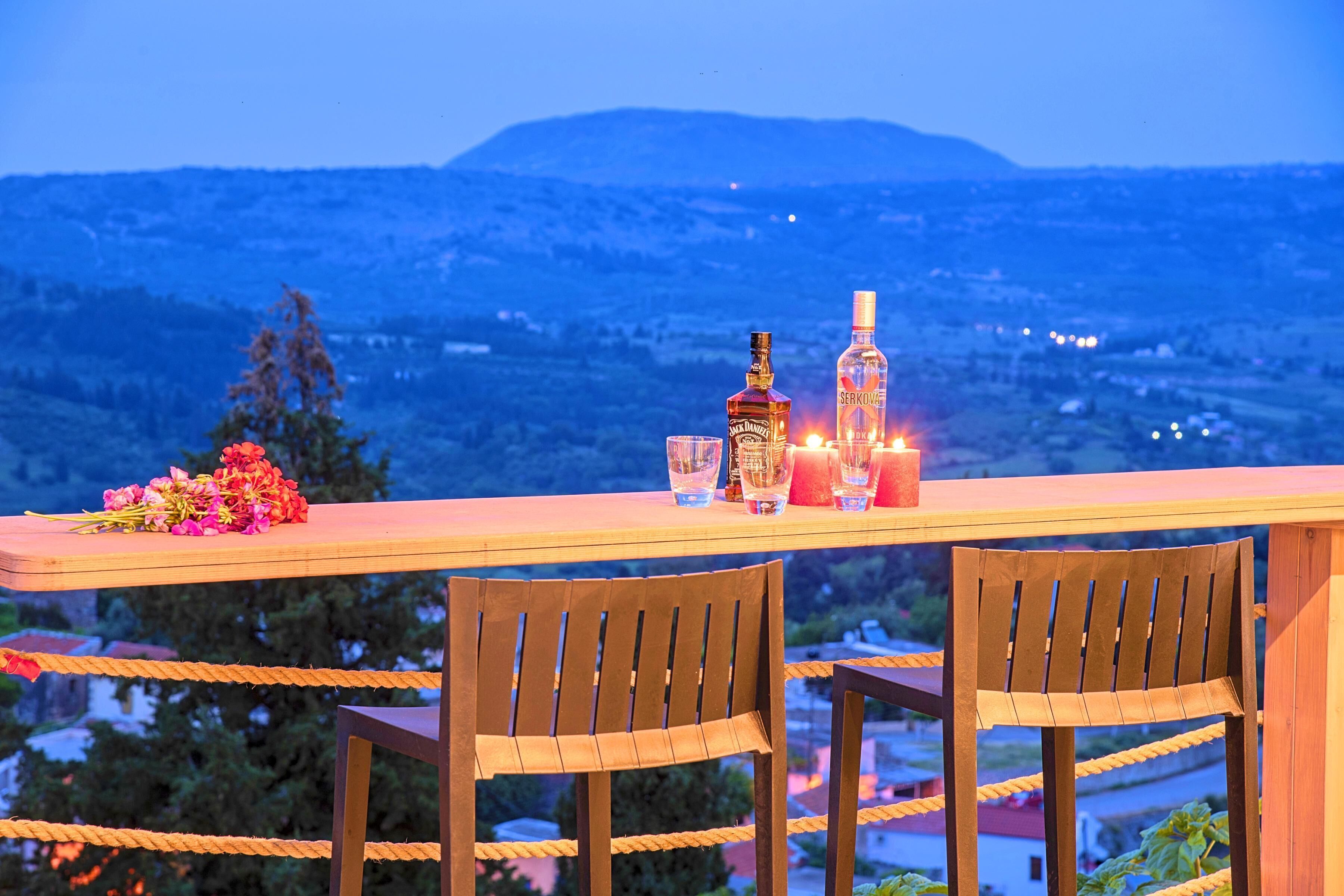 A table with a bottle, glasses, and a candle set on a balcony overlooking a mountainous landscape.