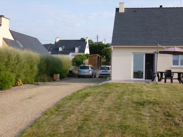 Une allée menant à une résidence, avec deux voitures garées, une pelouse et une maison dotée d’un toit en tuiles et d’une porte donnant sur un patio.