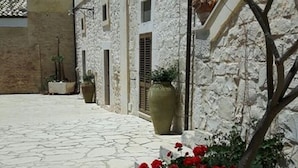 Une cour avec un sol en pierre, une grande plante en pot ornée de fleurs rouges et un bâtiment aux murs blancs et au toit recouvert de tuiles.