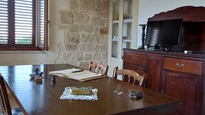 Un coin repas avec une grande table en bois, des chaises, un téléviseur posé sur un meuble en bois et un mur en pierre.