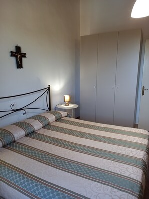 A bedroom with a bed, a bedside table with a lamp, a cross on the wall, and a wardrobe.