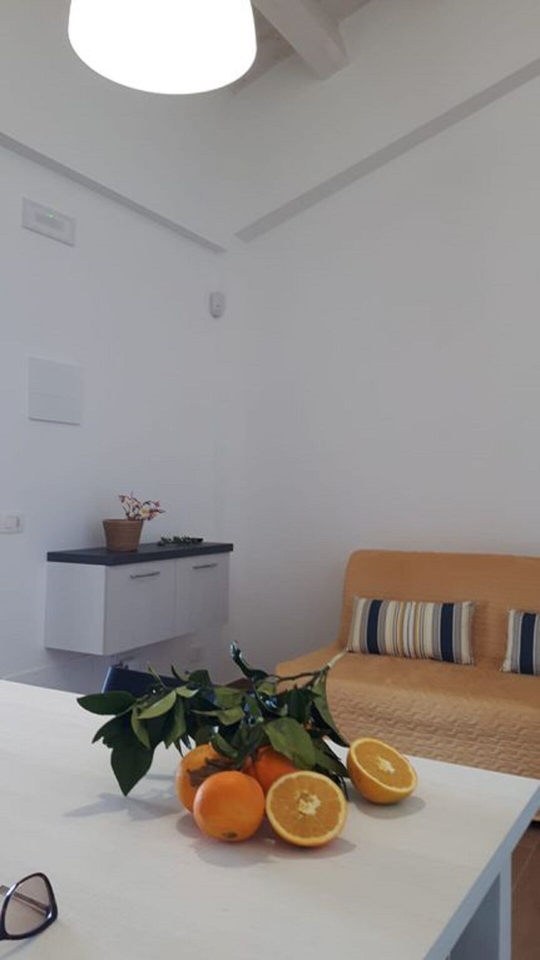 A modern living room with a white sofa, a dining table with oranges and a plant, and a kitchen in the background.