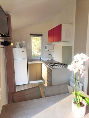 A compact kitchen with a refrigerator, a coffee maker, a microwave, and a stove. There is a window providing natural light, and a small potted orchid on the table. - Camping de La Grande Lande (Moelan-sur-Mer)