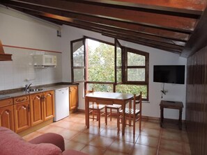 Una cocina con armarios de madera, un lavavajillas blanco, una mesa de comedor con sillas, una ventana con vistas a la vegetación y una mesita con un televisor en la pared.