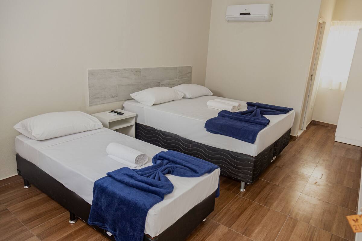 Two single beds with white linens and blue towels, a nightstand, and a wall-mounted air conditioner.