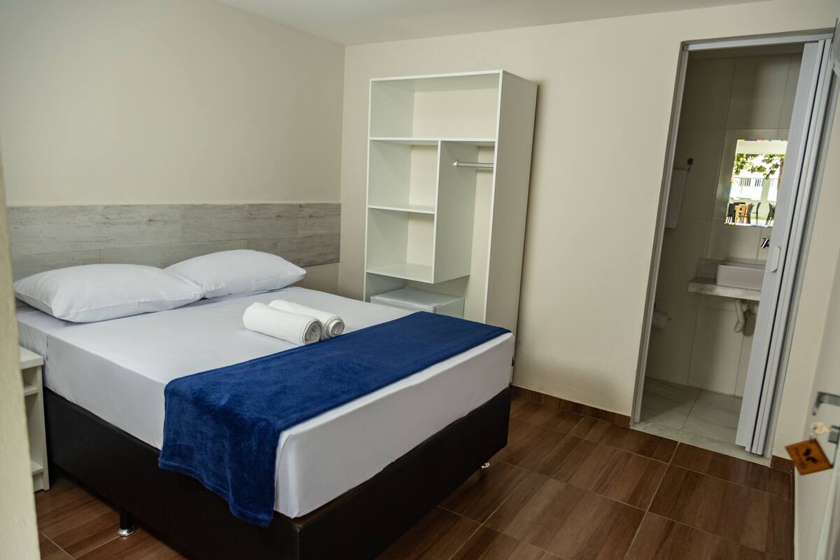 A bedroom with a bed, a blue blanket, a white pillow, a white bedside table, a white wardrobe, and a bathroom visible through an open door.