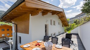 Una mesa de madera preparada para comer, con platos, cubiertos y vasos, y una cesta en un patio con vistas a una montaña.