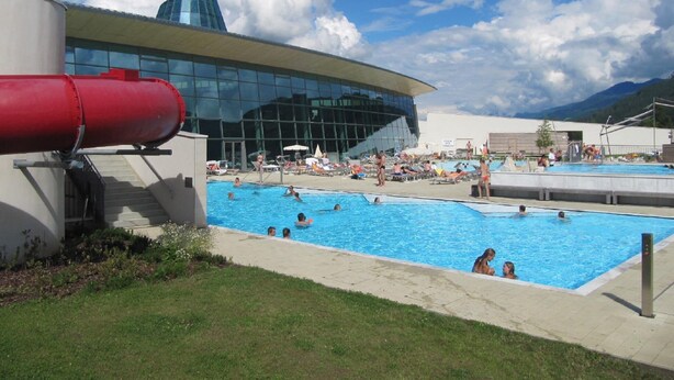 Zona de piscina con un edificio moderno al fondo, una tubería roja y gente disfrutando.