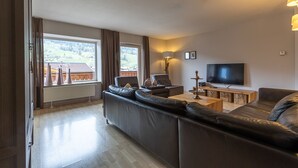 Una sala moderna con un sofá grande, una mesa de centro, un televisor y vistas a una cabaña de madera a través de un ventanal amplio.