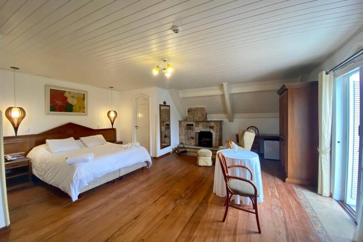Een ruime slaapkamer met houten vloer, een groot bed, een klein tafeltje met stoel en een open haard.