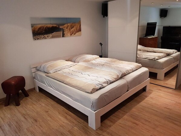 Ein Schlafzimmer mit einem Bett, einem kleinen Hocker, einem Gemälde und einem Spiegel, der ein Klavier und eine Kommode reflektiert.