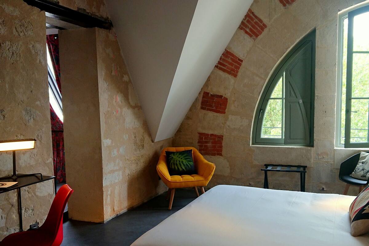 Een kamer met een bed, een bureau met een lamp, een gele stoel, een klein tafeltje en een raam met uitzicht op groen.
