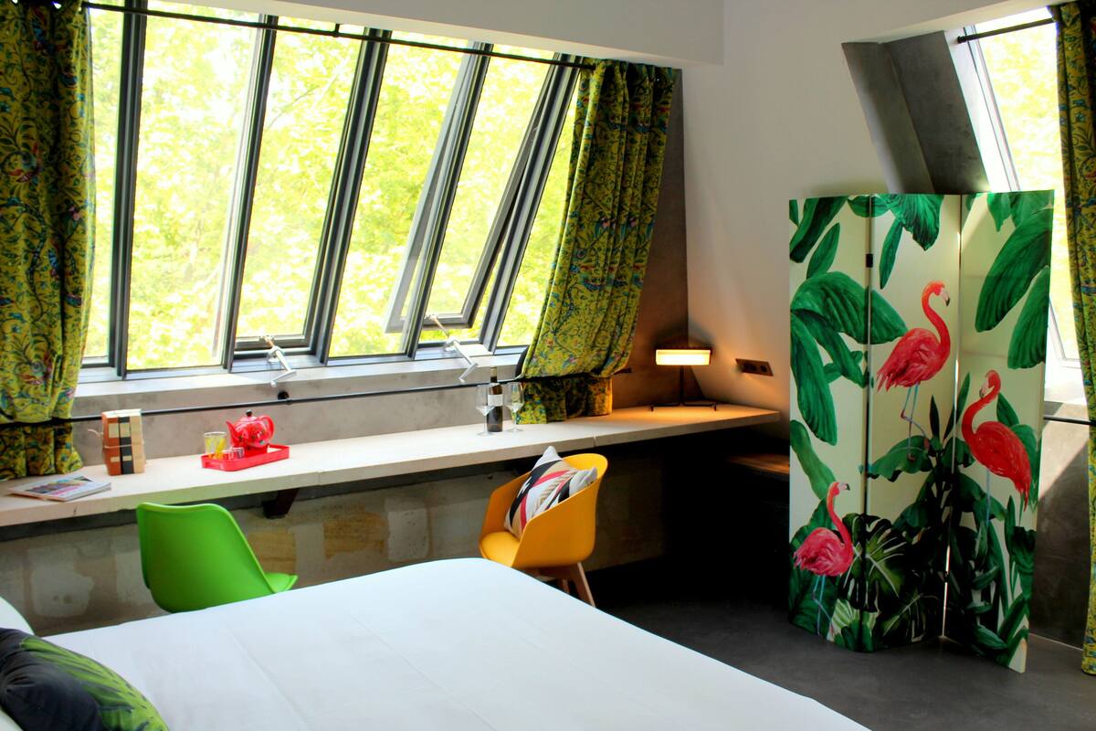 Een hotelkamer met een groot raam, een bureau met stoel, een bed en een decoratief paneel met flamingo's.