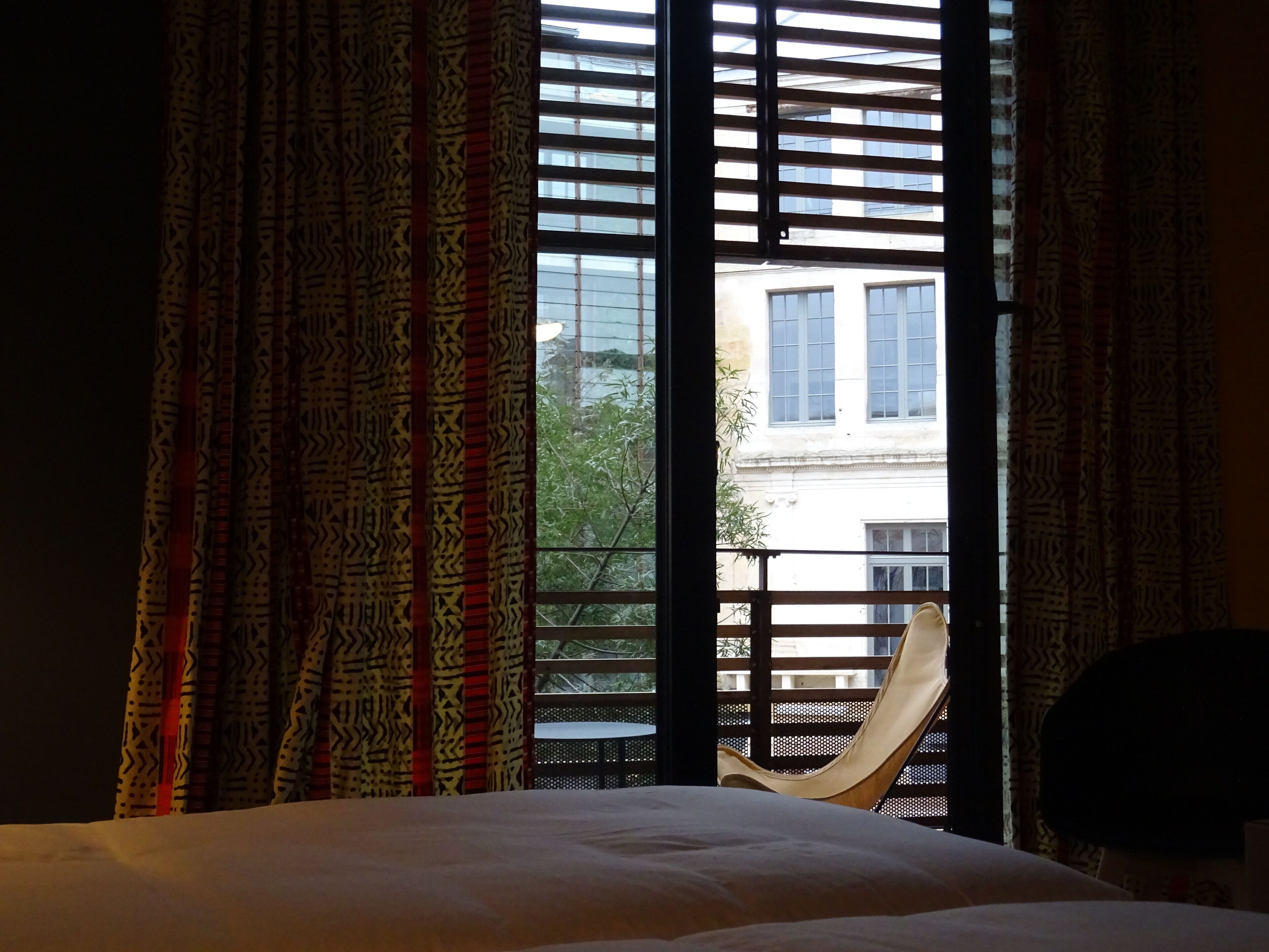 A hotel room with a bed, a chair, and a view of a building through a window with patterned curtains.