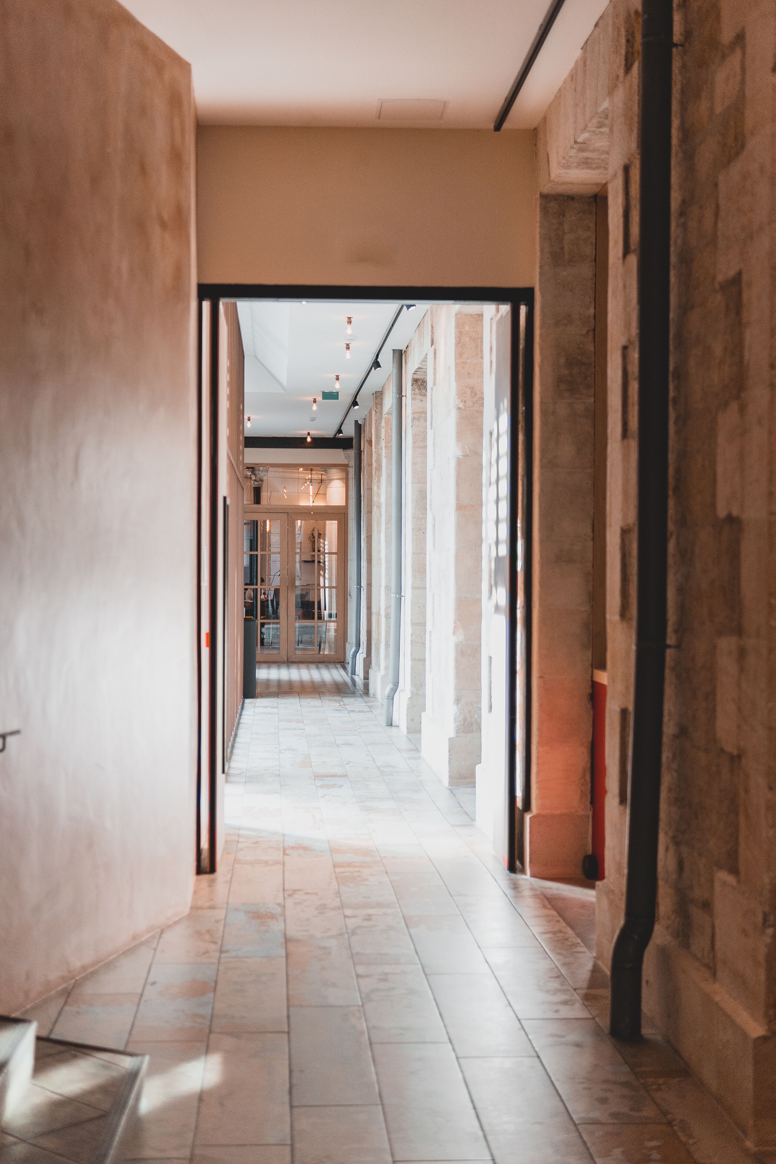 A corridor with stone walls and a tiled floor, leading to a glass door entrance.
