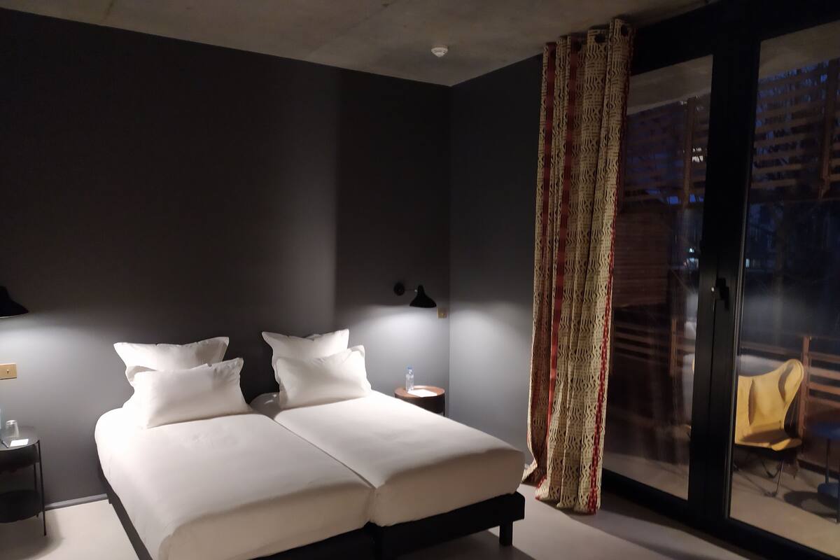 Een moderne hotelkamer met een groot bed, nachtkastjes, een kleintje tafeltje met een waterfles en uitzicht door een glazen deur.