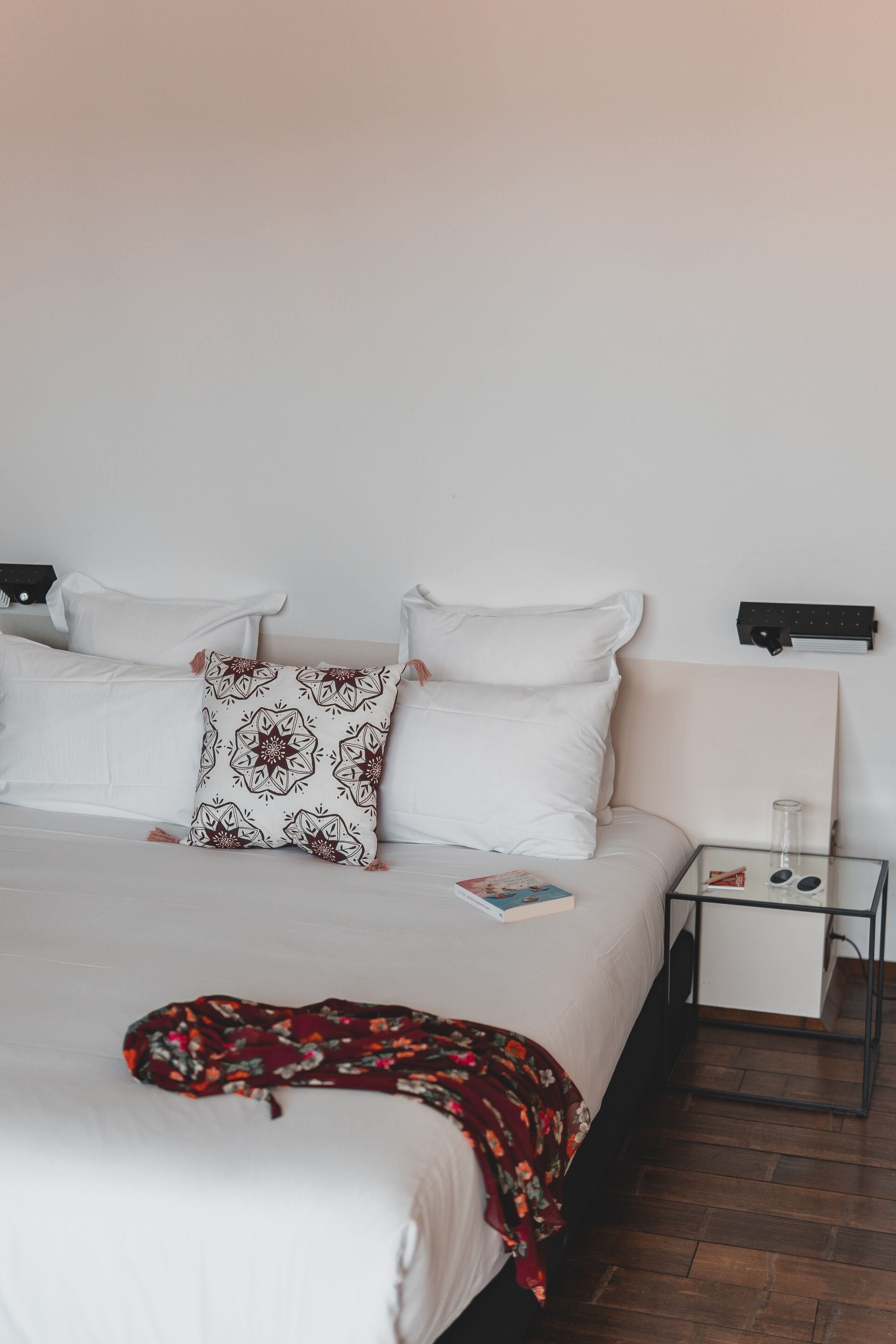 Un letto rifatto con lenzuola bianche e cuscini decorati, un comodino con una lampada e una coperta rossa sul pavimento.
