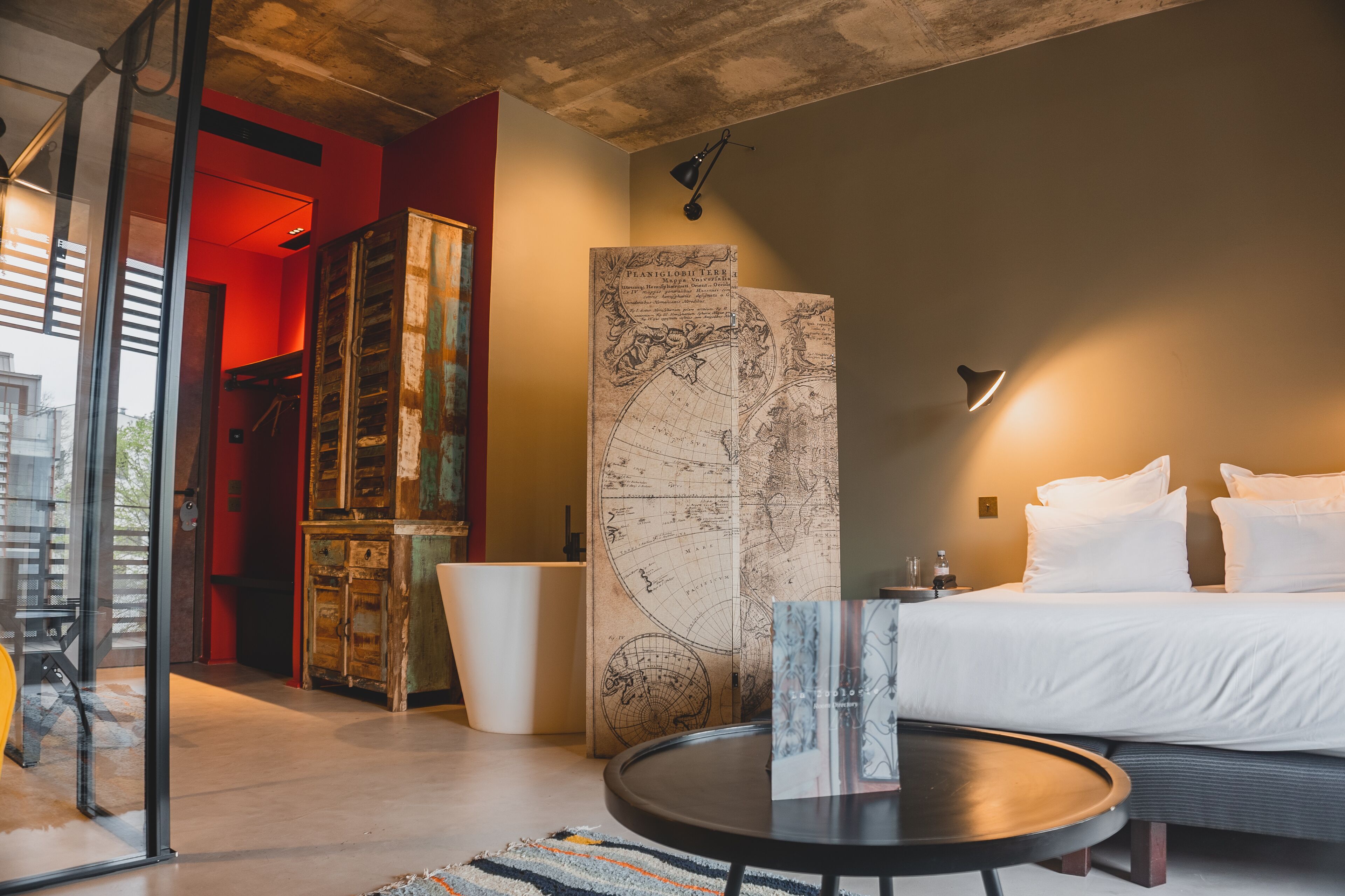 A modern hotel room with a large bed, a round table, a map on the wall, and a view of the city through a large window.