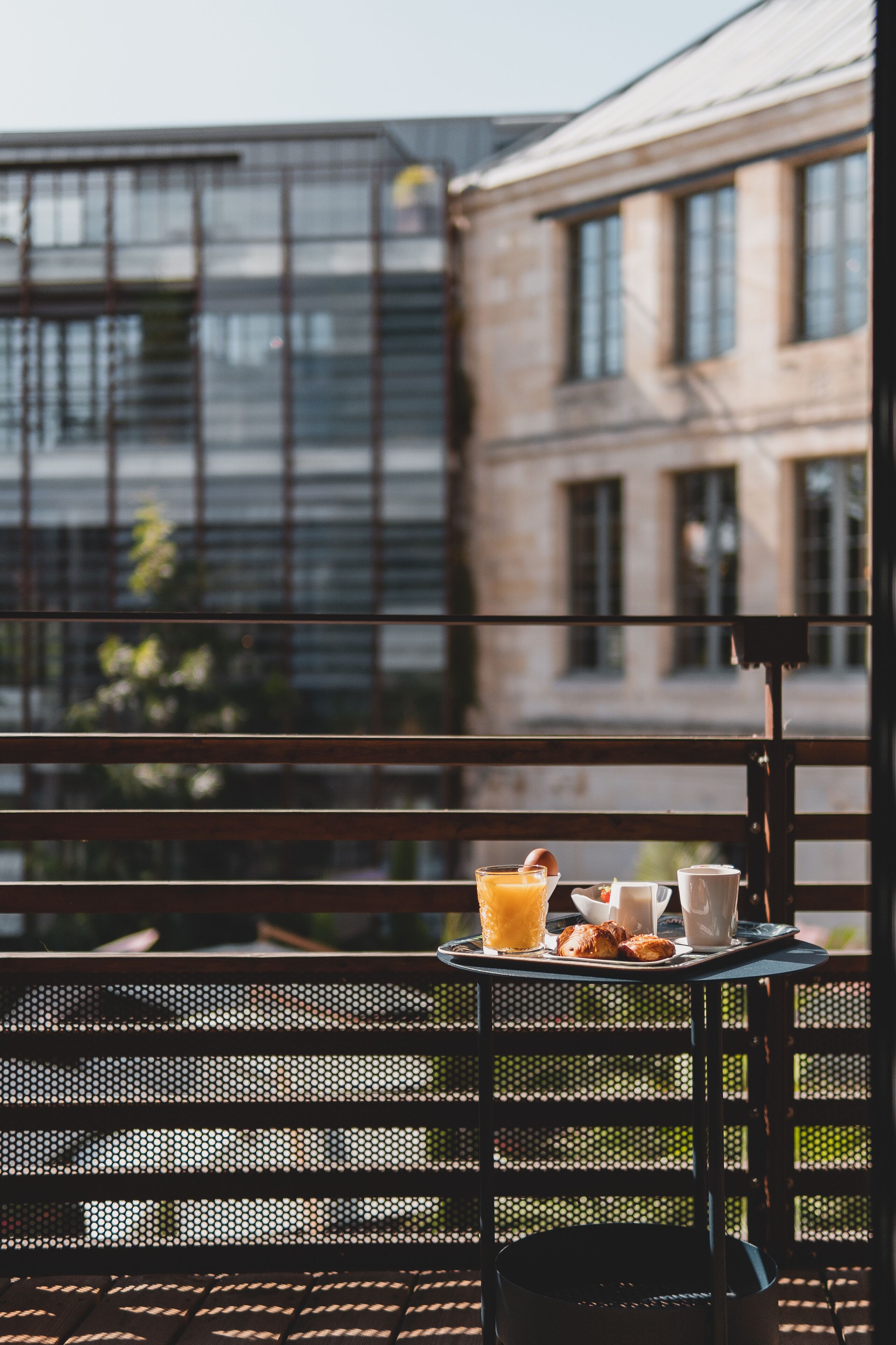 A metal table with a tray of breakfast items including orange juice, pastries, and a cup, set on a balcony with a view of a building and greenery.