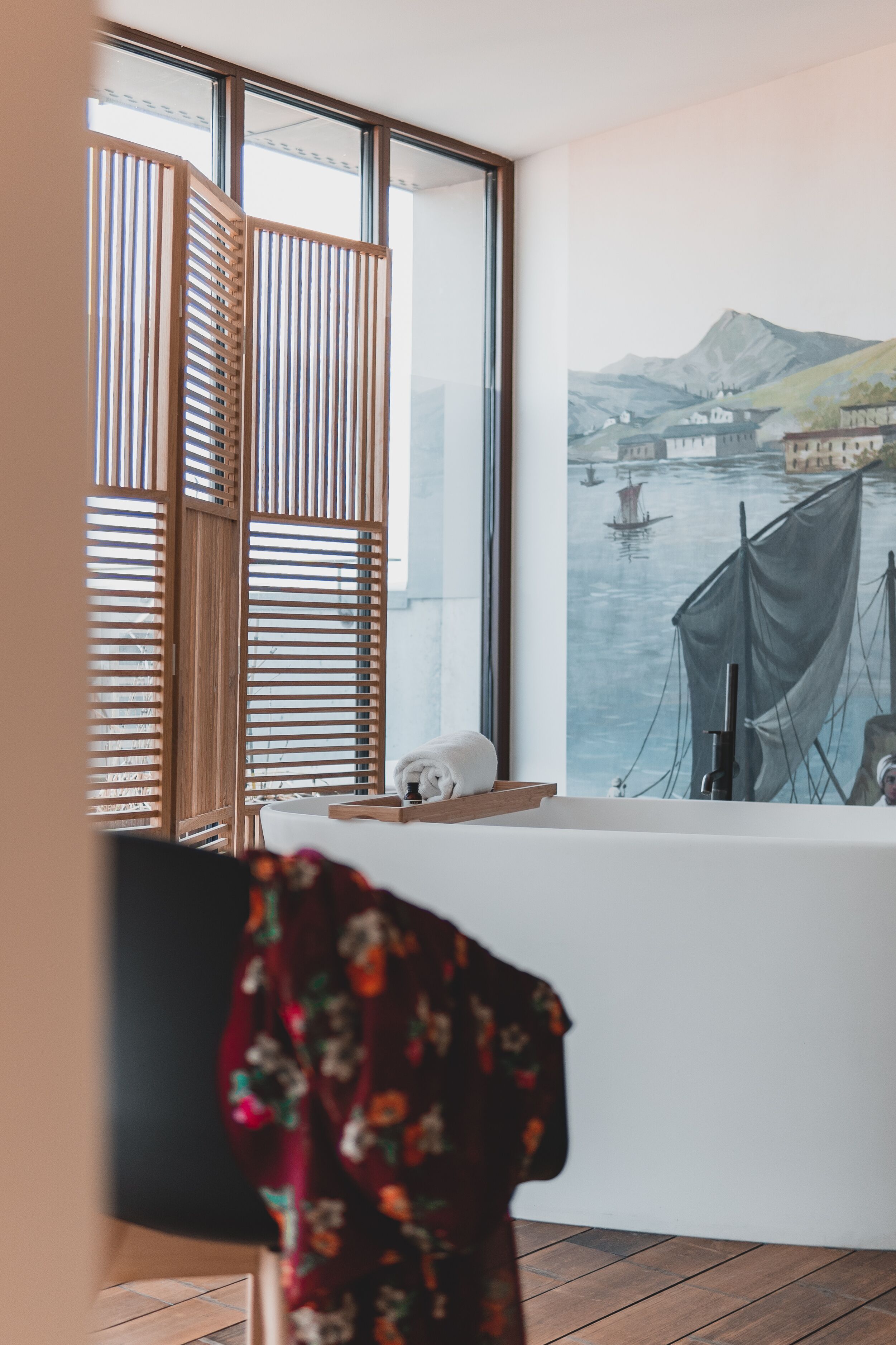 Una habitación con un ventanal grande, una persiana de listones de madera, una bañera blanca y un cuadro de una escena fluvial.