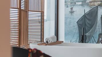 Una habitación con un ventanal grande, una persiana de listones de madera, una bañera blanca y un cuadro de una escena fluvial.