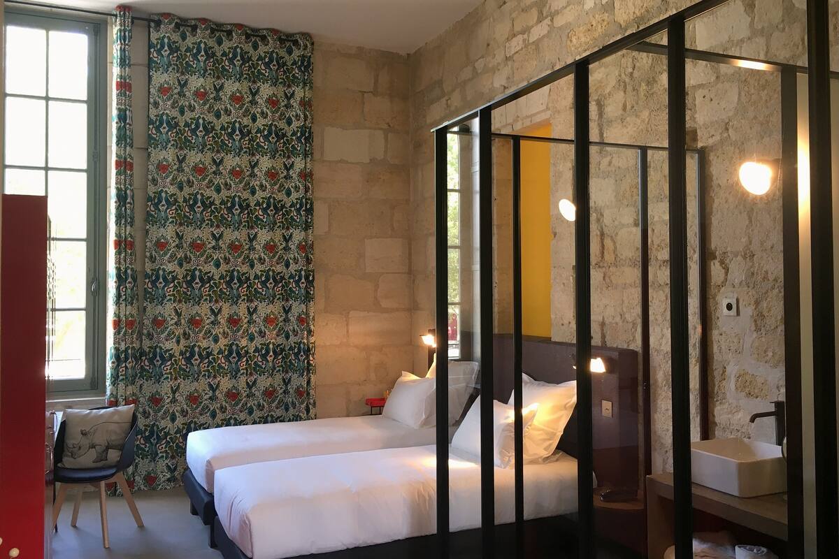 Een kamer met twee bedden, een stoel, een raam met gordijnen en een stenen muur.