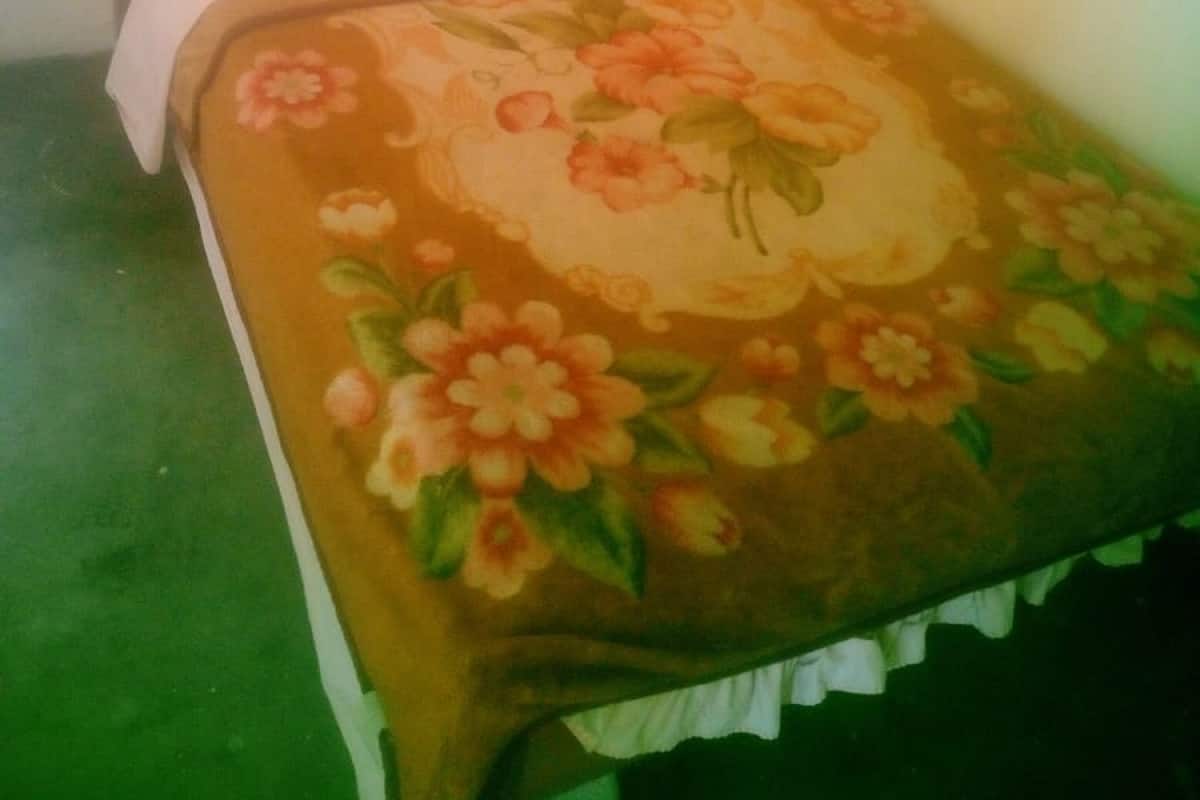 Una cama individual con colcha estampada y almohadas blancas en una habitación de paredes lisas y suelo verde.