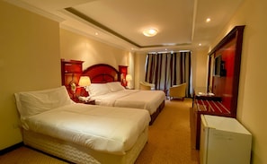 A hotel room with two beds, a desk, a chair, a TV, and a minibar. - New Penninsula Hotel (Dubai)