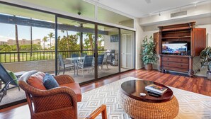 A spacious living area with a large sliding glass door leading to a patio, a comfortable seating area with a coffee table, and a television on a wooden cabinet. - Beautiful updates--everything new! Golf course views!  Huge 2/2 K. Royal #A301 (Lahaina)