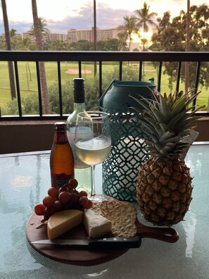 A table with a bottle of beer, a glass of white wine, a pineapple, grapes, cheese, crackers, and a knife on a balcony overlooking a golf course.