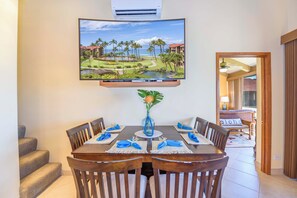 A dining area with a table set for six, a mounted TV displaying a tropical landscape, and a view of a pool and palm trees through an open door.