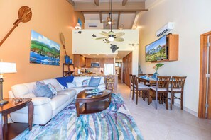 A spacious living area with a sofa, coffee table, dining table, and chairs. There is a ceiling fan, artwork on the walls, and an air conditioning unit.