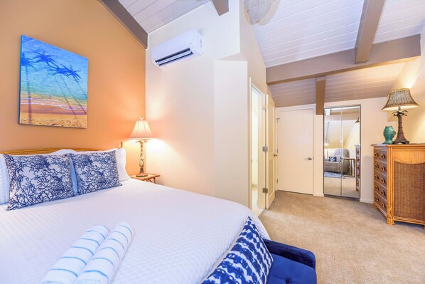 A neatly made bed with blue and white bedding, a wall-mounted air conditioner, and a beach-themed painting.